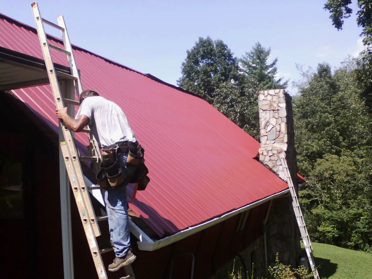 Roofer on a ladder beside a red metal roof and stone chimney — Soffit & Fascia Repair by Roofing Brothers in Terrell